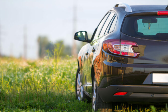 Back view of modern new shiny empty black car parked outside road in grassy looming field on bright summer sunny day on blurred green trees background. Transportation, pollution and ecology concept.