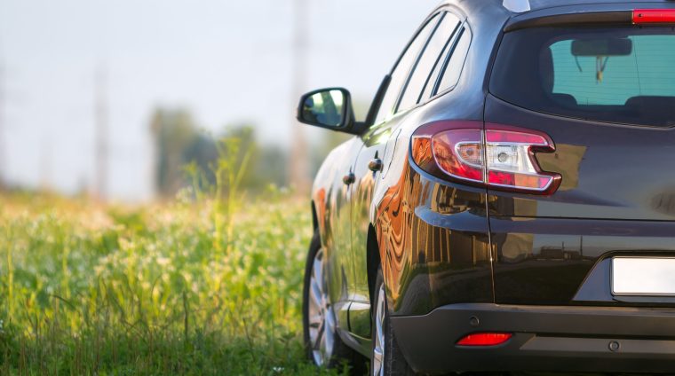 Back view of modern new shiny empty black car parked outside roa Back view of modern new shiny empty black car parked outside road in grassy looming field on bright summer sunny day on blurred green trees background. Transportation, pollution and ecology concept.